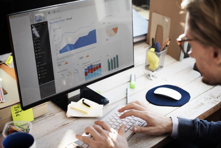 Pessoa trabalhando em computador, analisando gráficos e dados de desempenho exibidos na tela em um ambiente de escritório.