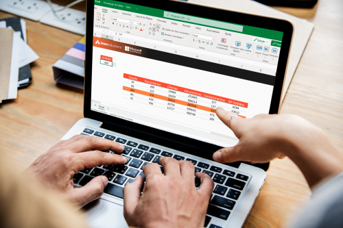 Mãos de duas pessoas interagindo em frente a um notebook exibindo planilha do Excel com formatação condicional destacando dados em vermelho.